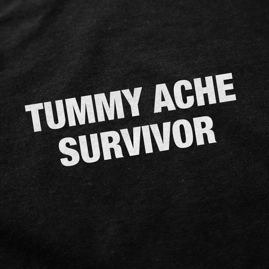 White text on black fabric reads TUMMY ACHE SURVIVOR in bold, slanted letters, making the Tummy Ache Survivor T-Shirt a perfect blend of humor and comfort for anyone who loves funny shirts.