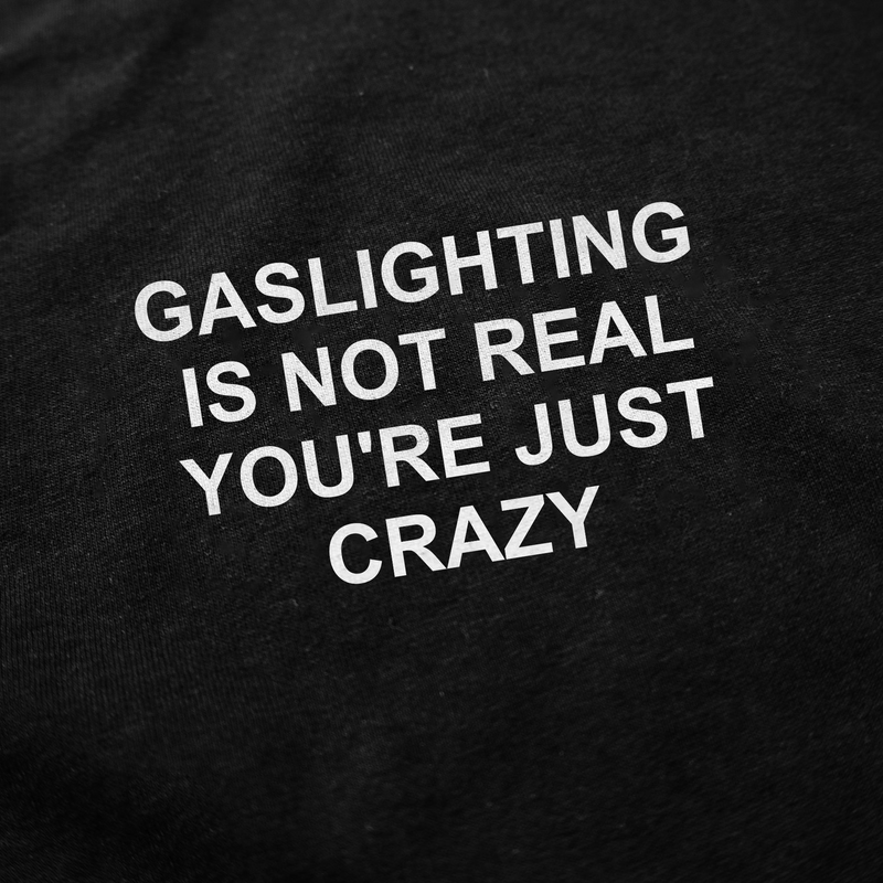 The Gaslighting Is Not Real You're Just Crazy T-Shirt features bold white text on black fabric that reads: “Gaslighting is not real you’re just crazy.”.