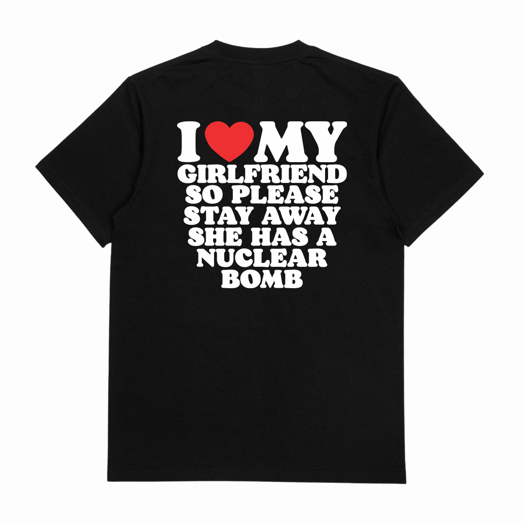 The "I Heart My Girlfriend So Please Stay Away She Has A Nuclear Bomb T-Shirt (Backprint)" is a black cotton tee made from ethically sourced cotton, with bold white text and a red heart symbol on the back.