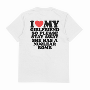 The "I Heart My Girlfriend So Please Stay Away She Has A Nuclear Bomb T-Shirt (Backprint)" is a white cotton tee with ethically grown fibers, featuring bold black text and a red heart graphic on the back.