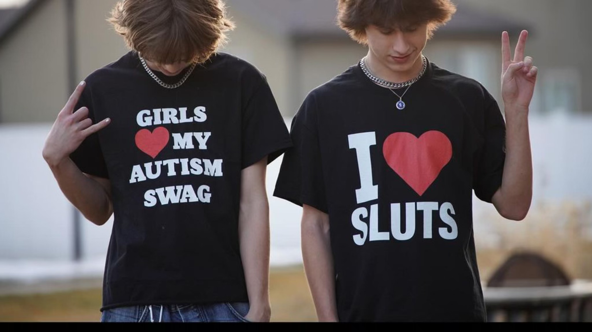 Two young people stand side by side outdoors, both wearing black t-shirts with bold white and red text. One shirt reads Girls ❤️ my autism swag, and the other says I ❤️ sluts. Both are making hand gestures.