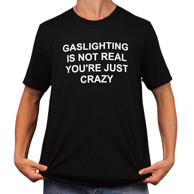 A person holds the sides of a Gaslighting Is Not Real You're Just Crazy T-Shirt, featuring the bold white phrase "GASLIGHTING IS NOT REAL YOURE JUST CRAZY"—perfect for fans of edgy and Illegal Shirts humor.